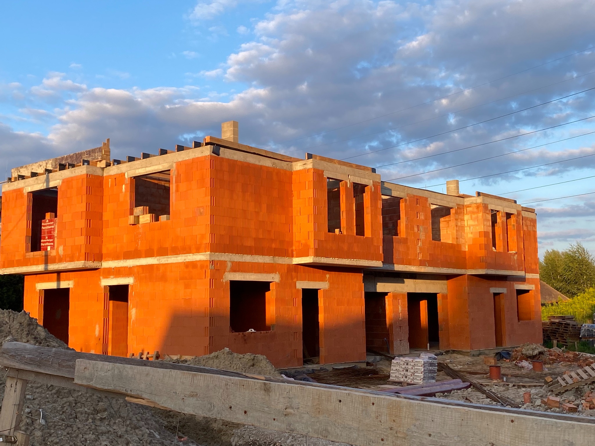 Surowy stan dwukondygnacyjnego budynku z czerwonej cegły ceramicznej w trakcie budowy, z widocznymi otworami okiennymi i drzwiowymi, na tle błękitnego nieba z obłokami.