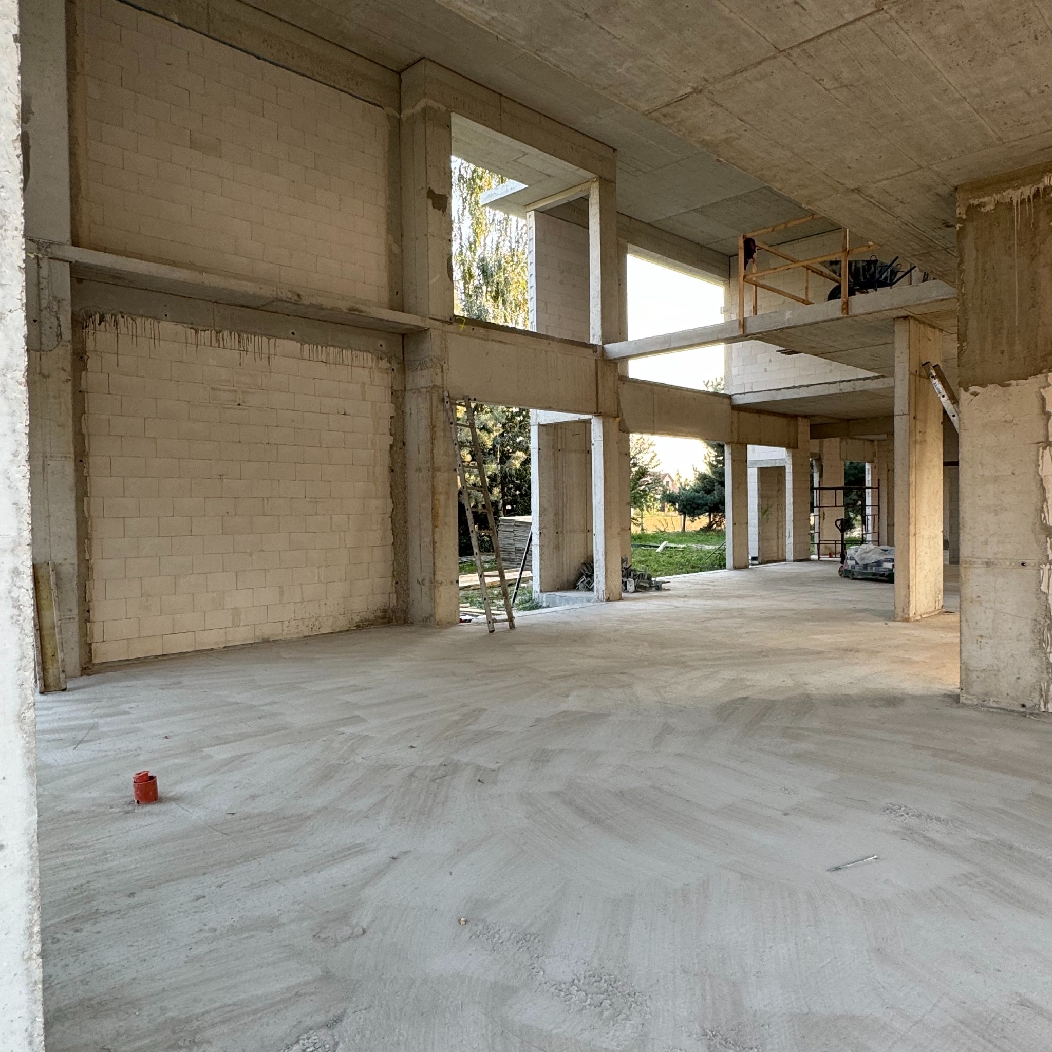 Wnętrze surowego stanu budynku z betonu komórkowego i betonu, widoczne surowe ściany z pustaków, betonowe słupy i strop, drabina oparta o ścianę, na zewnątrz zieleń.