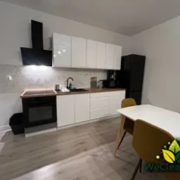 Wnętrze czystej, nowocześnie urządzonej kuchni z białymi szafkami, czarnym sprzętem AGD i drewnianą podłogą. Na pierwszym planie biały stół jadalny z krzesłami w kolorze musztardowym. W prawym...