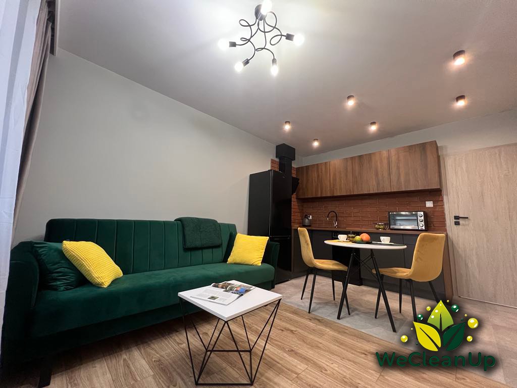 Wnętrze salonu z zieloną sofą i żółtymi poduszkami, widok na kuchnię z ceglaną ścianą, okrągły stół z owocami i logo firmy sprzątającej w prawym dolnym rogu.