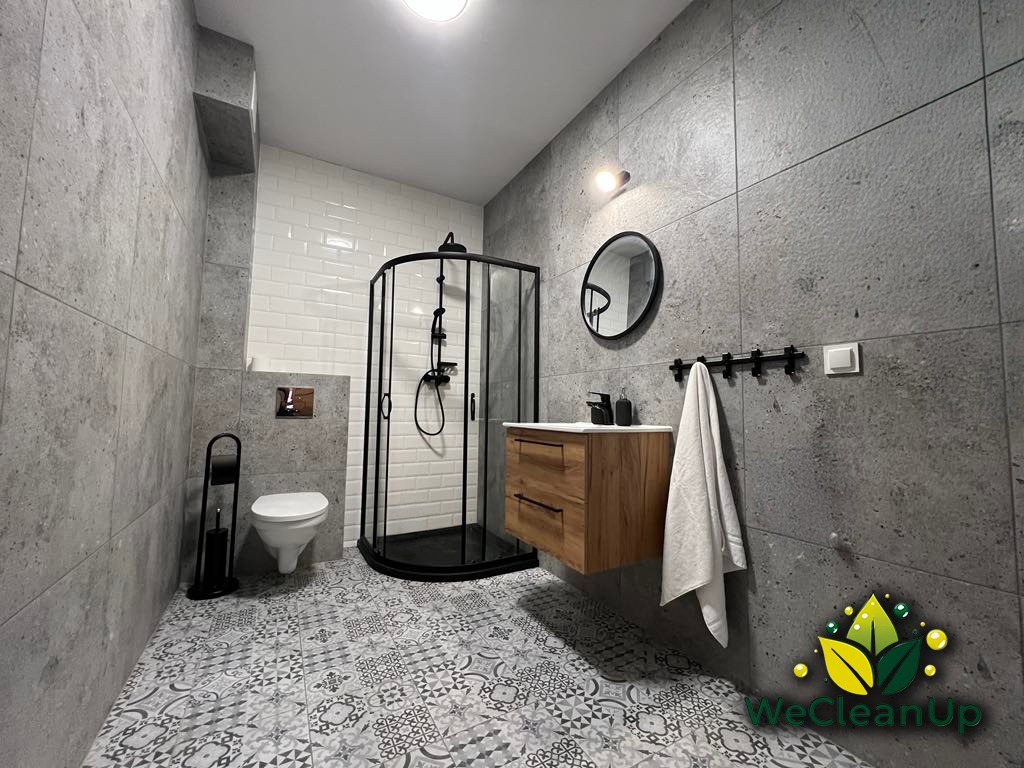 Nowoczesna łazienka z szarymi płytkami, czarną kabiną prysznicową narożną, drewnianą szafką pod umywalką i ozdobną podłogą w stylu retro. Widoczne logo firmy sprzątającej w prawym dolnym rogu.