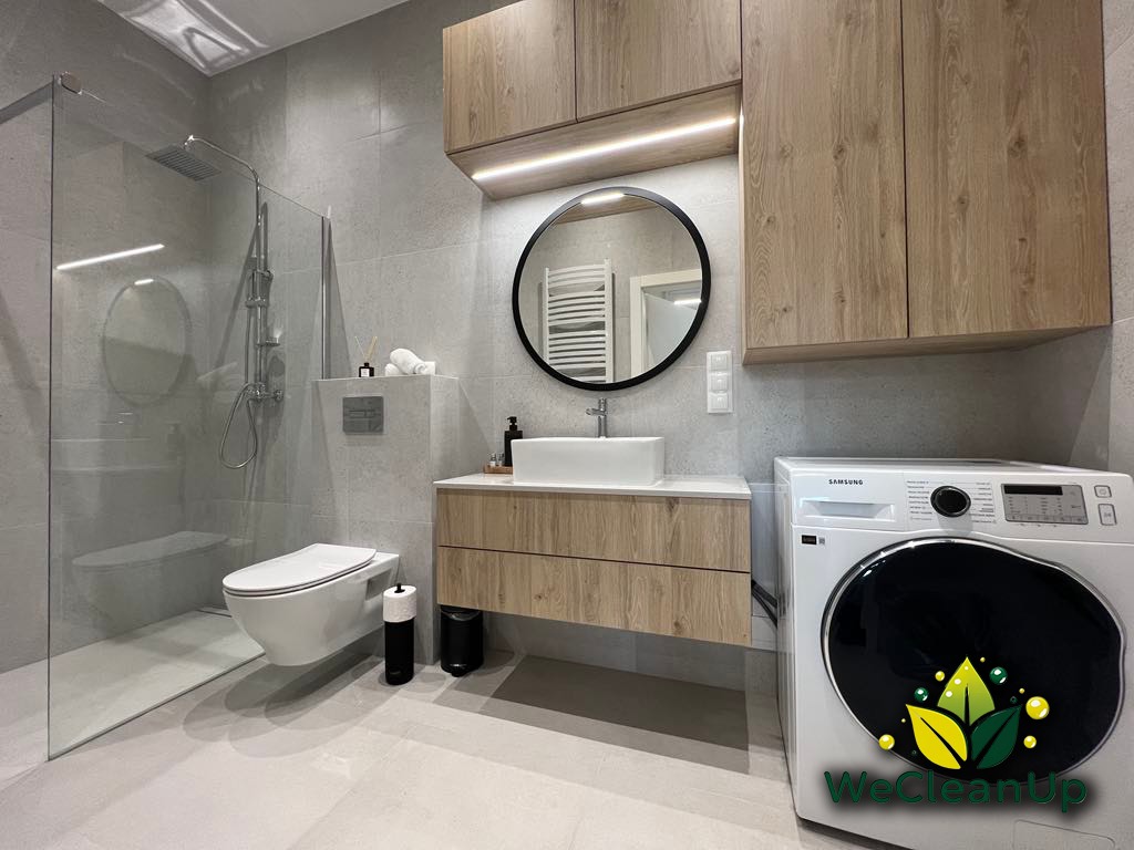 Nowoczesna łazienka w odcieniach szarości z drewnianymi szafkami, kabiną prysznicową typu walk-in, umywalką nablatową i pralką Samsung z logo WeCleanup na drzwiczkach.