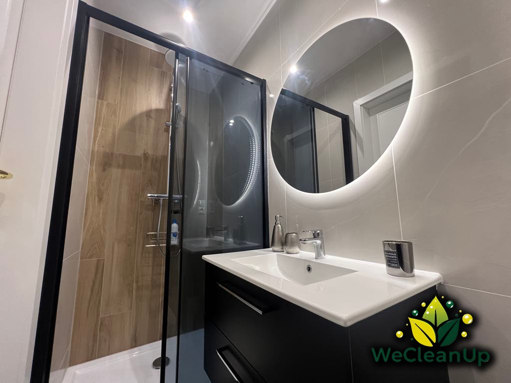 Nowoczesna łazienka po sprzątaniu: kabina prysznicowa z czarną ramą, okrągłe lustro z podświetleniem LED, umywalka z czarną szafką, szare płytki imitujące marmur, widoczne logo firmy sprzątającej...