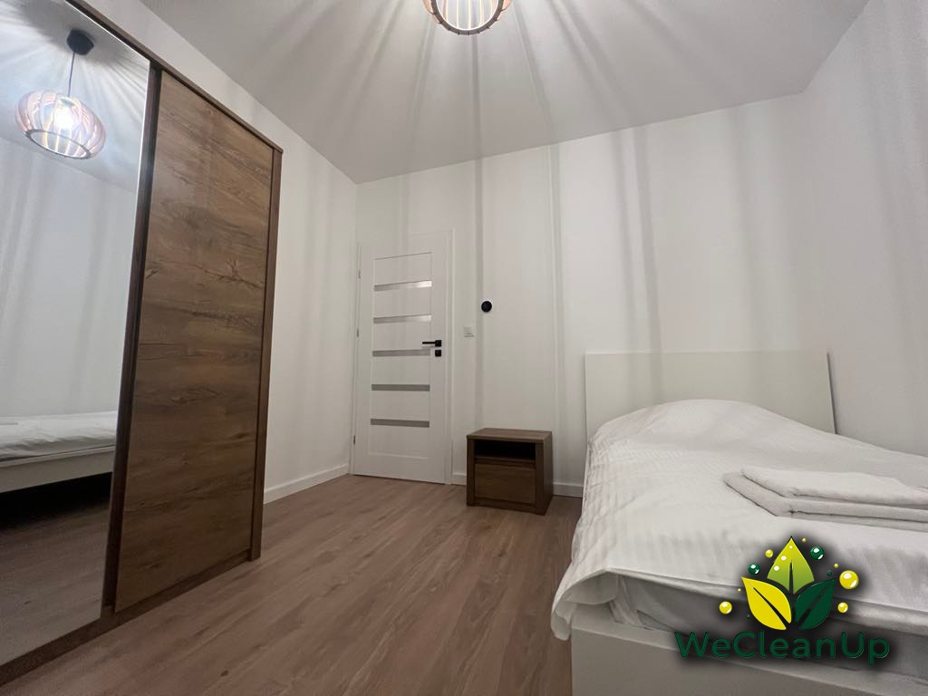 Wnętrze czystej sypialni z łóżkiem pojedynczym, szafką nocną, lustrzaną szafą przesuwną i drewnianą podłogą, oświetlone lampą sufitową. Na kołdrze leżą złożone białe ręczniki. W prawym dolnym rogu...