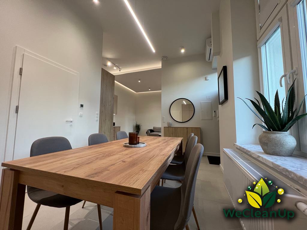 Jasne wnętrze jadalni z drewnianym stołem i szarymi krzesłami, widok na salon, roślina doniczkowa na parapecie, logo firmy sprzątającej w prawym dolnym rogu.