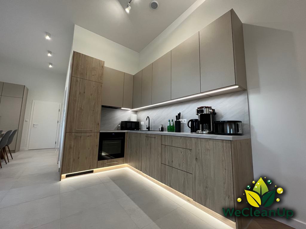 Nowoczesna kuchnia z beżowo-szarymi szafkami, podświetleniem LED pod szafkami i na podłodze, oraz logo firmy sprzątającej w prawym dolnym rogu.