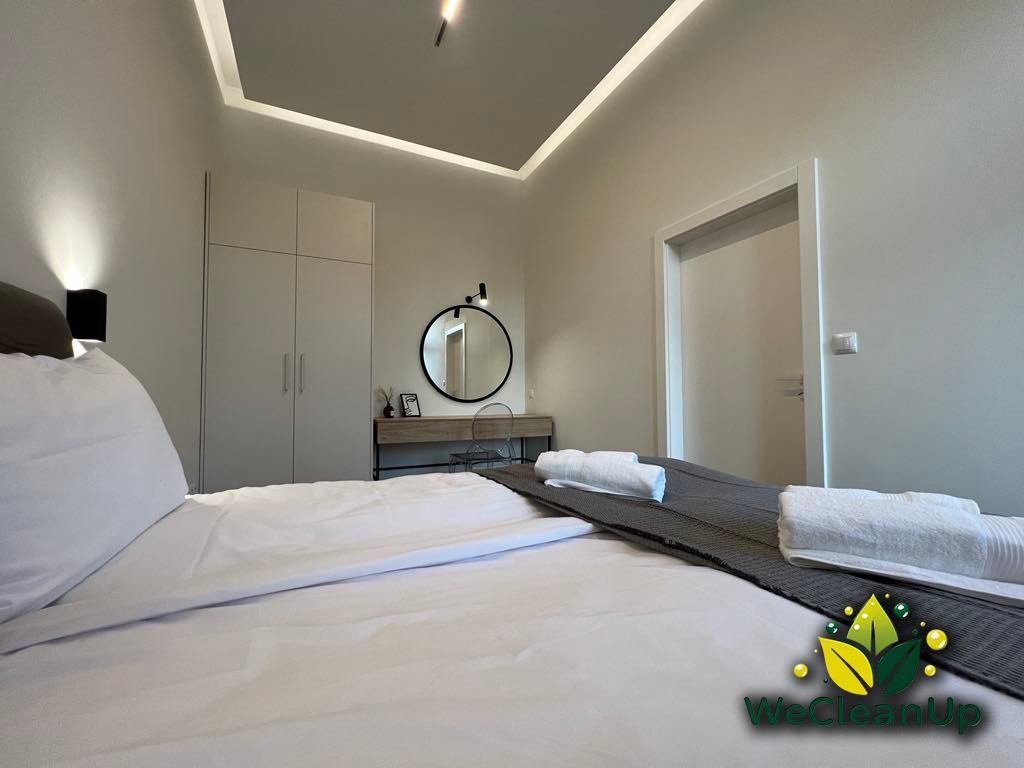 Jasne wnętrze sypialni z łóżkiem w bieli, szafą w zabudowie i toaletką z okrągłym lustrem, widoczne ręczniki i logo WeCleanUp w prawym dolnym rogu.