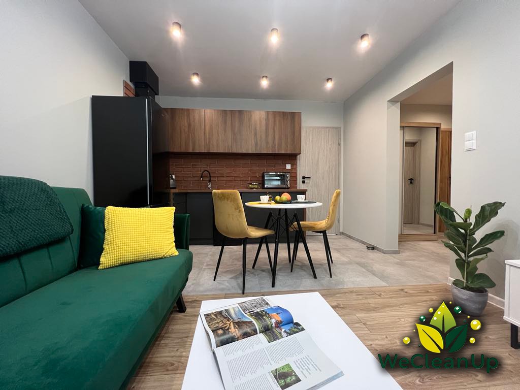 Jasne, nowocześnie urządzone wnętrze mieszkania z zieloną sofą, żółtą poduszką, okrągłym stołem z owocami, otwartą książką na białym stoliku kawowym i rośliną doniczkową w rogu, po sprzątaniu.