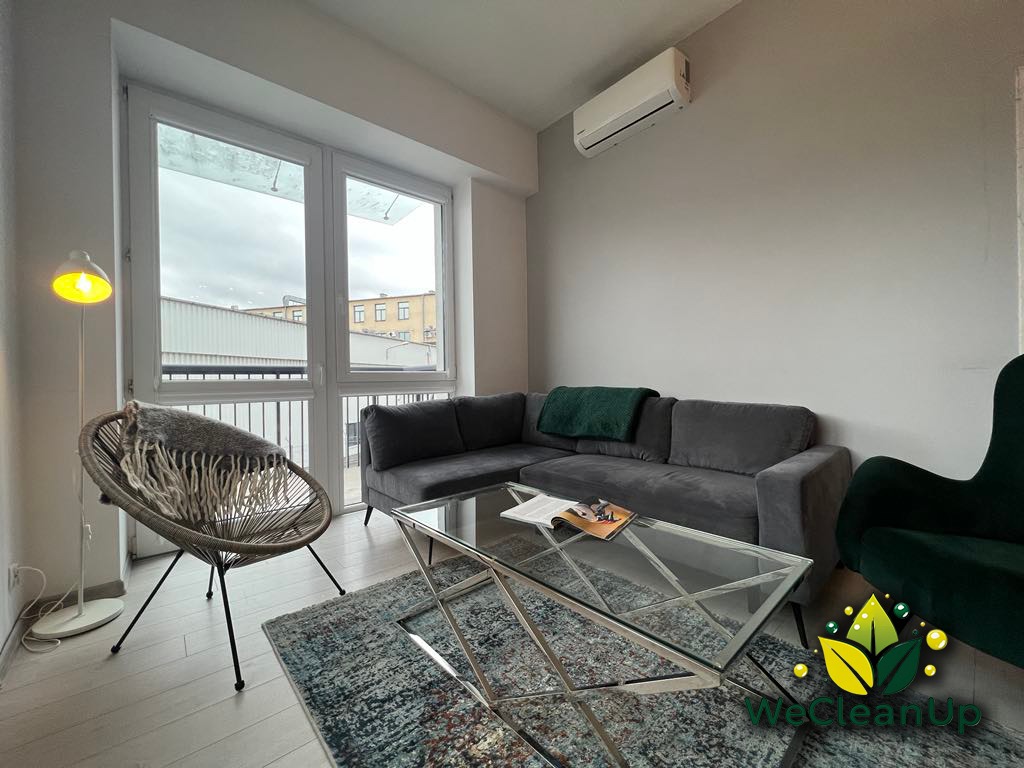Uporządkowany salon z szarą sofą narożną, szklanym stolikiem kawowym i wiklinowym fotelem z pledem, widok na okno z widocznym budynkiem przemysłowym w tle, logo firmy WeCleanUp w prawym dolnym rogu.