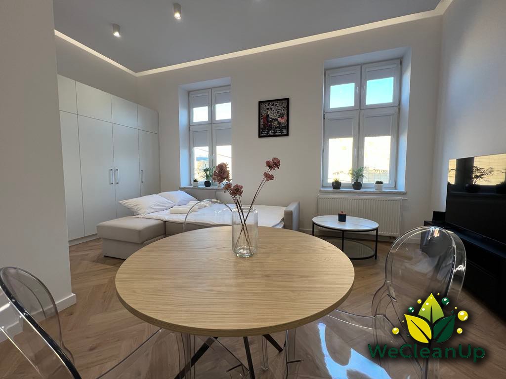 Jasne, nowocześnie urządzone wnętrze mieszkania z drewnianym stołem na pierwszym planie, kanapą z białymi ręcznikami w tle i logo firmy sprzątającej WeCleanUp na przezroczystym krześle.
