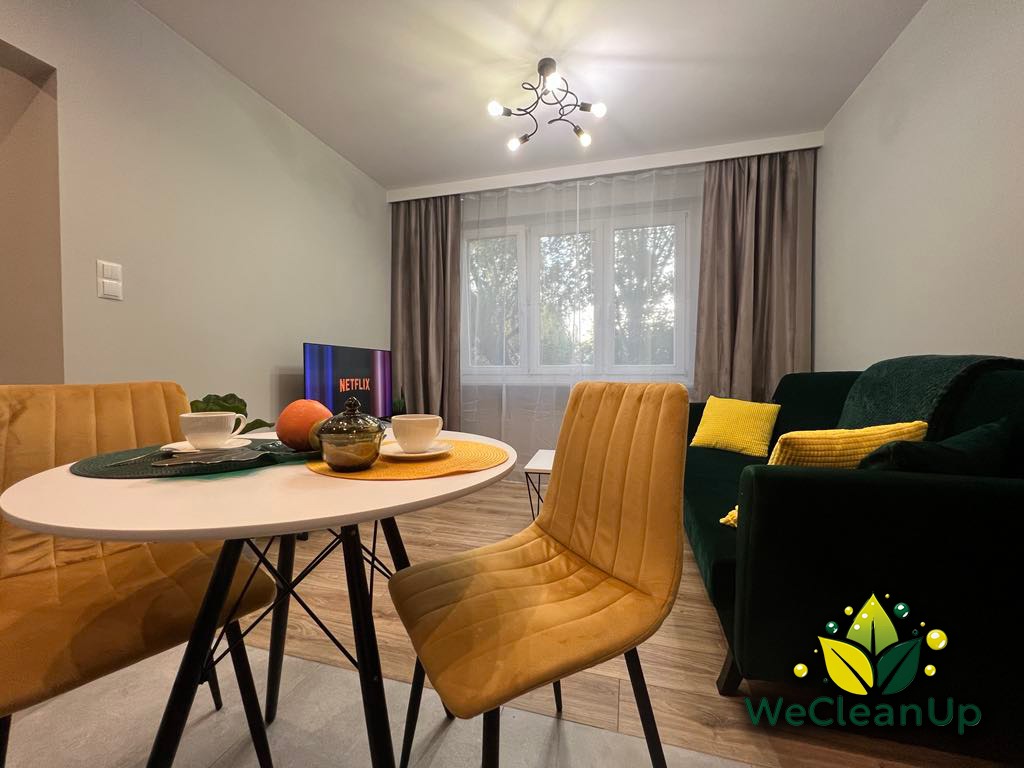 Salon z ciemnozieloną sofą, żółtymi poduszkami i krzesłami, okrągłym stołem z filiżankami i owocami, telewizorem z logo Netflix oraz logo firmy sprzątającej WeCleanUp w prawym dolnym rogu.