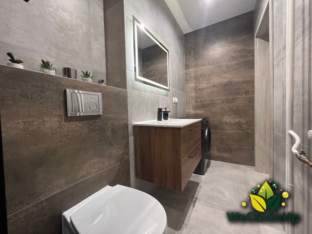 Nowoczesna łazienka z szarymi i brązowymi płytkami, podświetlanym lustrem, drewnianą szafką pod umywalką i pralką w tle, z widocznym logo firmy sprzątającej w prawym dolnym rogu.