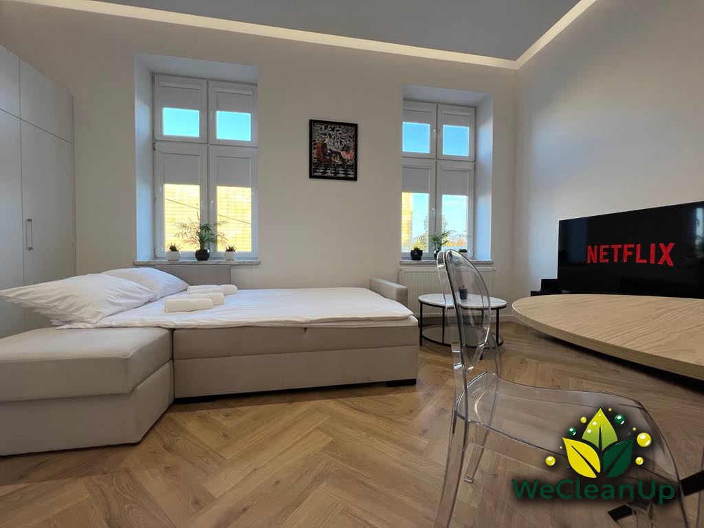 Jasne, nowocześnie urządzone wnętrze mieszkania w Rzeszowie, z łóżkiem kontynentalnym, telewizorem i parkietem ułożonym w jodełkę. Na parapecie rośliny w doniczkach.