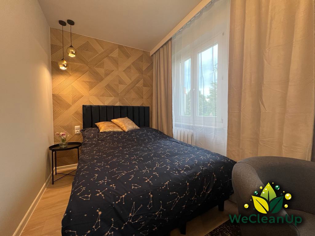 Uporządkowana sypialnia z łóżkiem w pościeli w gwiazdy, dekoracyjną ścianą i lampami wiszącymi, widoczne logo firmy sprzątającej WeCleanUp na fotelu.
