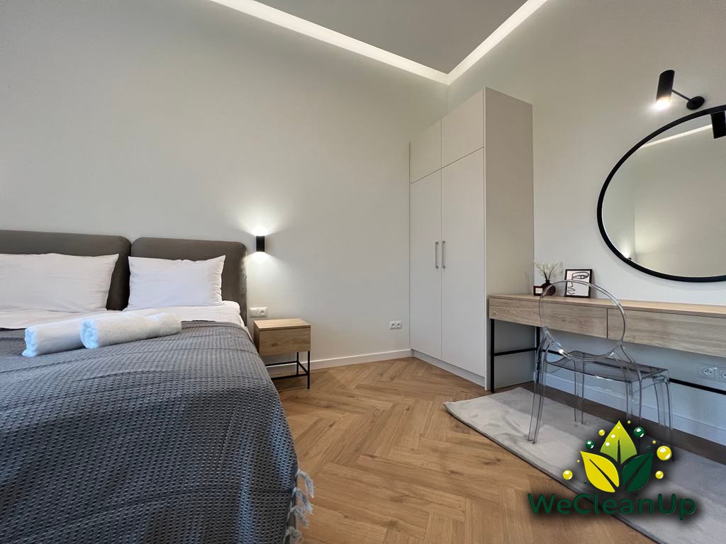 Jasna sypialnia po sprzątaniu: zaścielone łóżko z szarą narzutą i białymi ręcznikami, parkiet w jodełkę, biała szafa i toaletka z okrągłym lustrem.