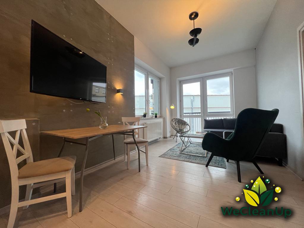 Jasne wnętrze mieszkania z drewnianą podłogą, szarą ścianą z telewizorem, stołem z kwiatem w wazonie, krzesłami i sofą w tle, ozdobione logo firmy sprzątającej w rogu.