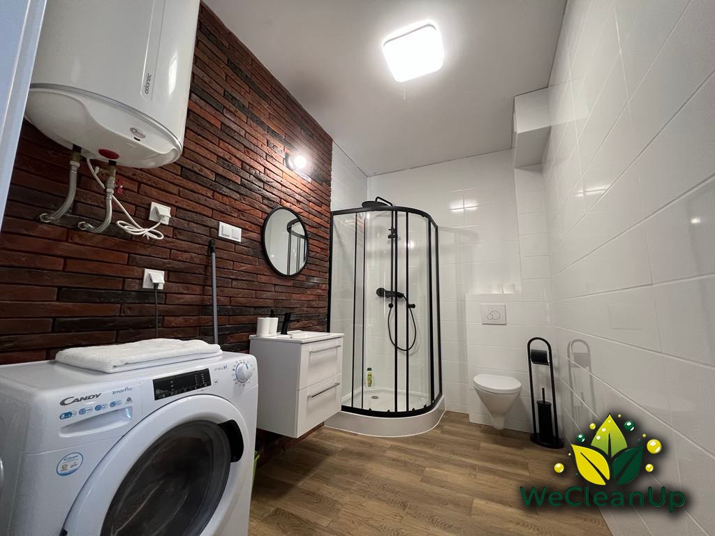 Nowoczesna łazienka z pralką Candy SmartPro, kabiną prysznicową z czarną ramą, toaletą, umywalką z szafką i ścianą z cegły, po sprzątaniu, widoczne logo firmy sprzątającej w prawym dolnym rogu.