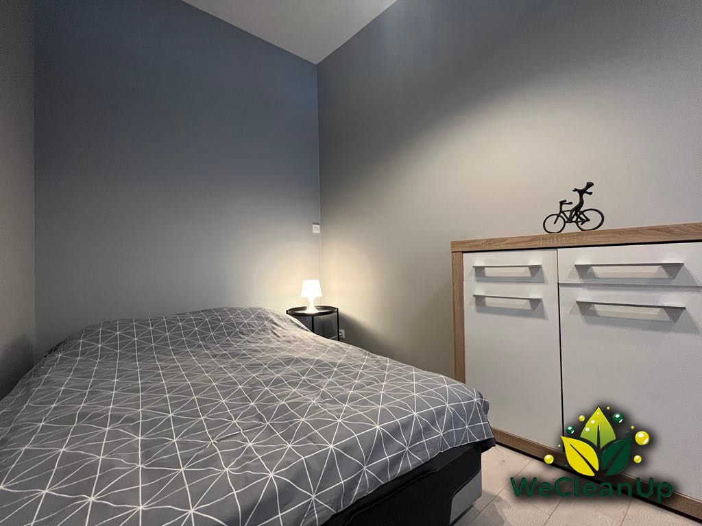 Pokój po posprzątaniu: łóżko z szarą pościelą w geometryczne wzory, biała szafka z drewnianym blatem, dekoracja roweru, lampka nocna, logo firmy sprzątającej w prawym dolnym rogu.