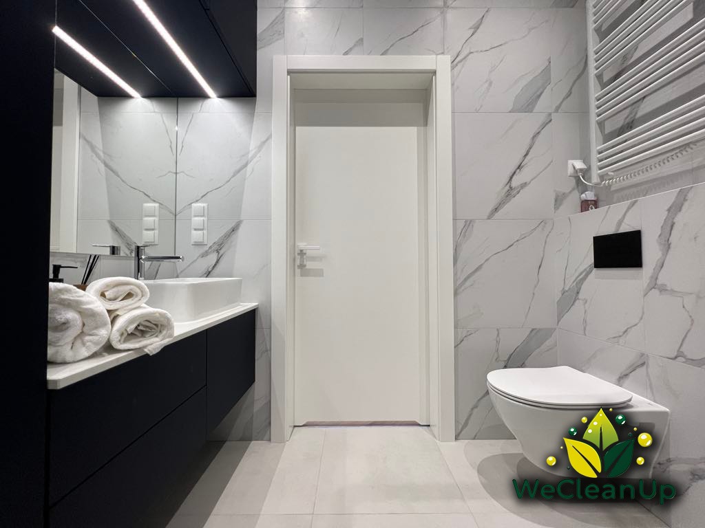 Nowoczesna łazienka z marmurowymi płytkami, czarną szafką pod umywalką, białymi ręcznikami i toaletą wiszącą; widoczne oświetlenie LED na suficie.