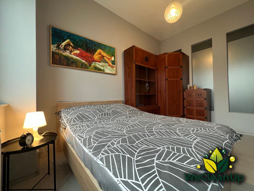 Uporządkowana sypialnia z łóżkiem w geometryczną pościel, drewnianymi meblami i obrazem nad wezgłowiem, widoczne logo firmy sprzątającej w rogu.