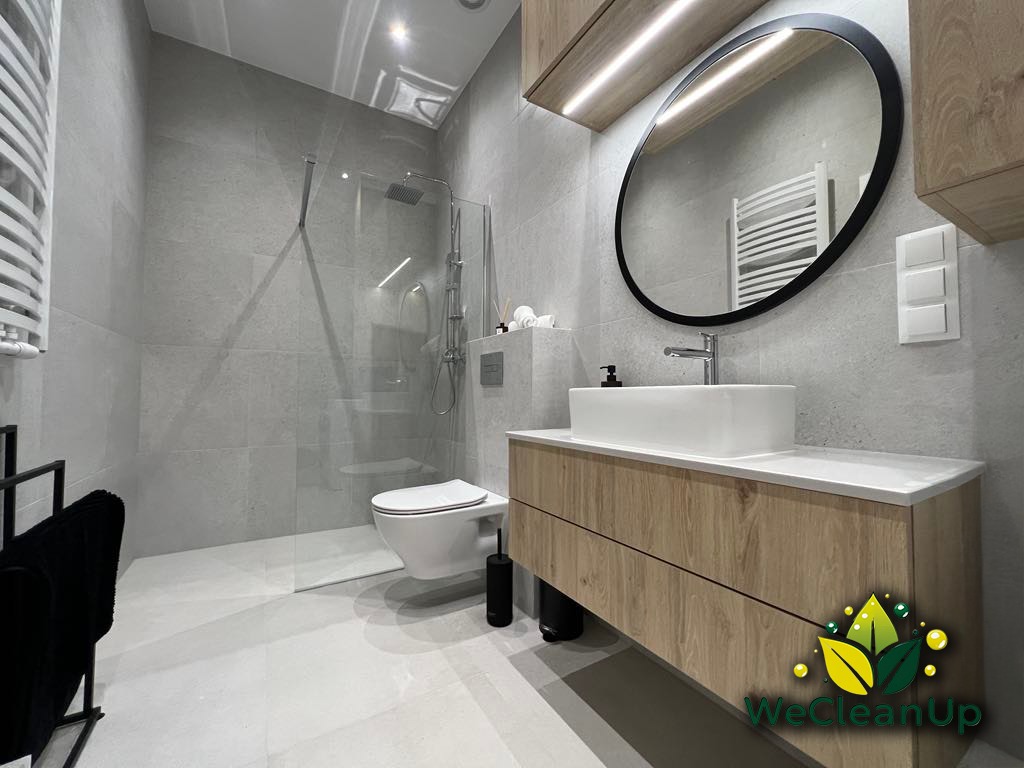 Nowoczesna, wyczyszczona łazienka z kabiną prysznicową typu walk-in, umywalką nablatową na drewnianej szafce i okrągłym lustrem, widoczny logo firmy WeCleanUp.