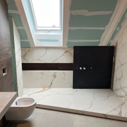 Avenue Refinish - Łazienka w trakcie remontu: białe marmurowe płytki, czarna ściana prysznicowa, podwieszana toaleta, okno dachowe i płyty karton-gips na suficie. Widoczne elementy montażowe.