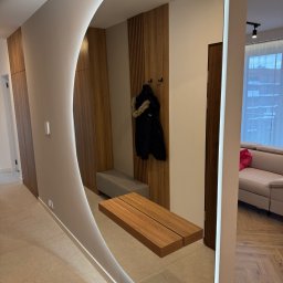 Avenue Refinish - Nowoczesny przedpokój z owalnym lustrem z podświetleniem LED, drewnianą zabudową i siedziskiem. Widoczna podłoga z płytek i fragment salonu z sofą.