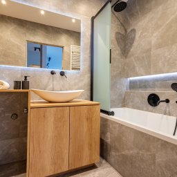 Avenue Refinish - Nowoczesna łazienka z marmurowymi płytkami, drewnianą szafką pod umywalką i czarną armaturą. Oświetlenie LED podkreśla detale. Roślina w doniczce dodaje świeżości.