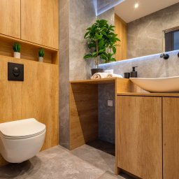 Avenue Refinish - Nowoczesna łazienka z drewnianymi szafkami, szarą płytką imitującą marmur i owalną umywalką. Uwagę zwraca wisząca toaleta i roślina doniczkowa na blacie.