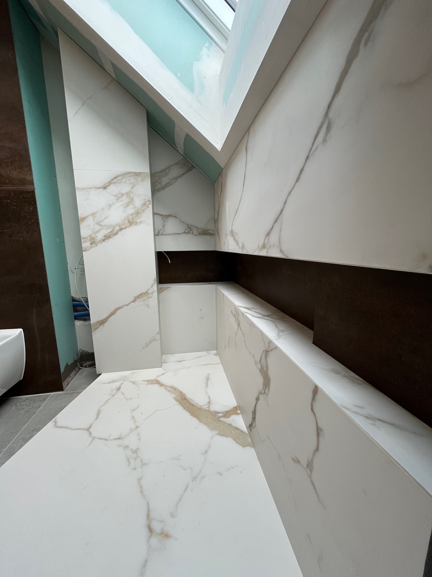 Nowoczesna łazienka w trakcie wykańczania: białe płytki imitujące marmur z brązowymi żyłami, zabudowa wnęki z półkami, widoczne elementy instalacji.