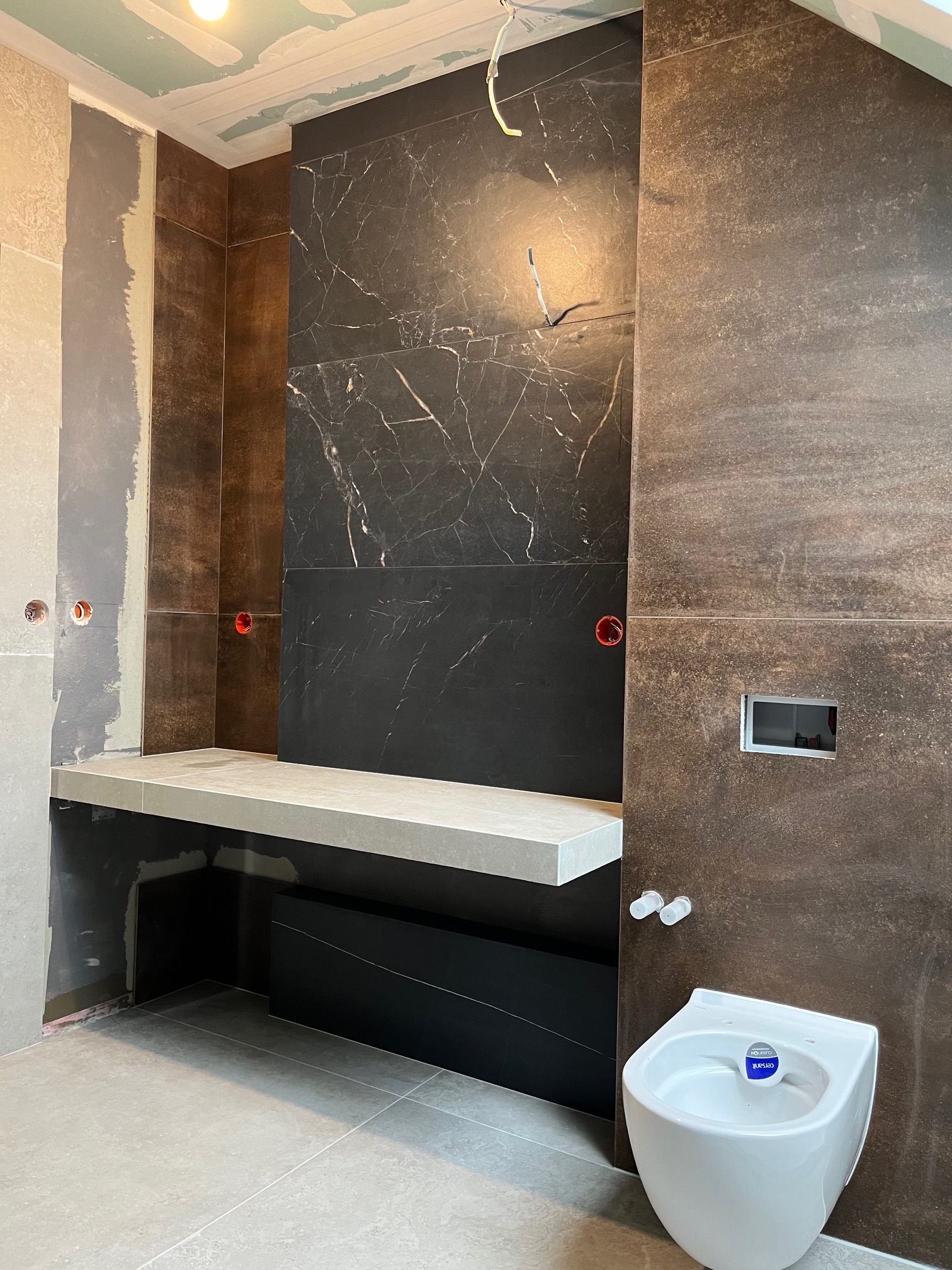 Wnętrze łazienki w trakcie wykańczania: brązowe i czarne płytki, białe bidet i umywalka, szara półka. Widoczne elementy instalacji elektrycznej i sanitarnej.