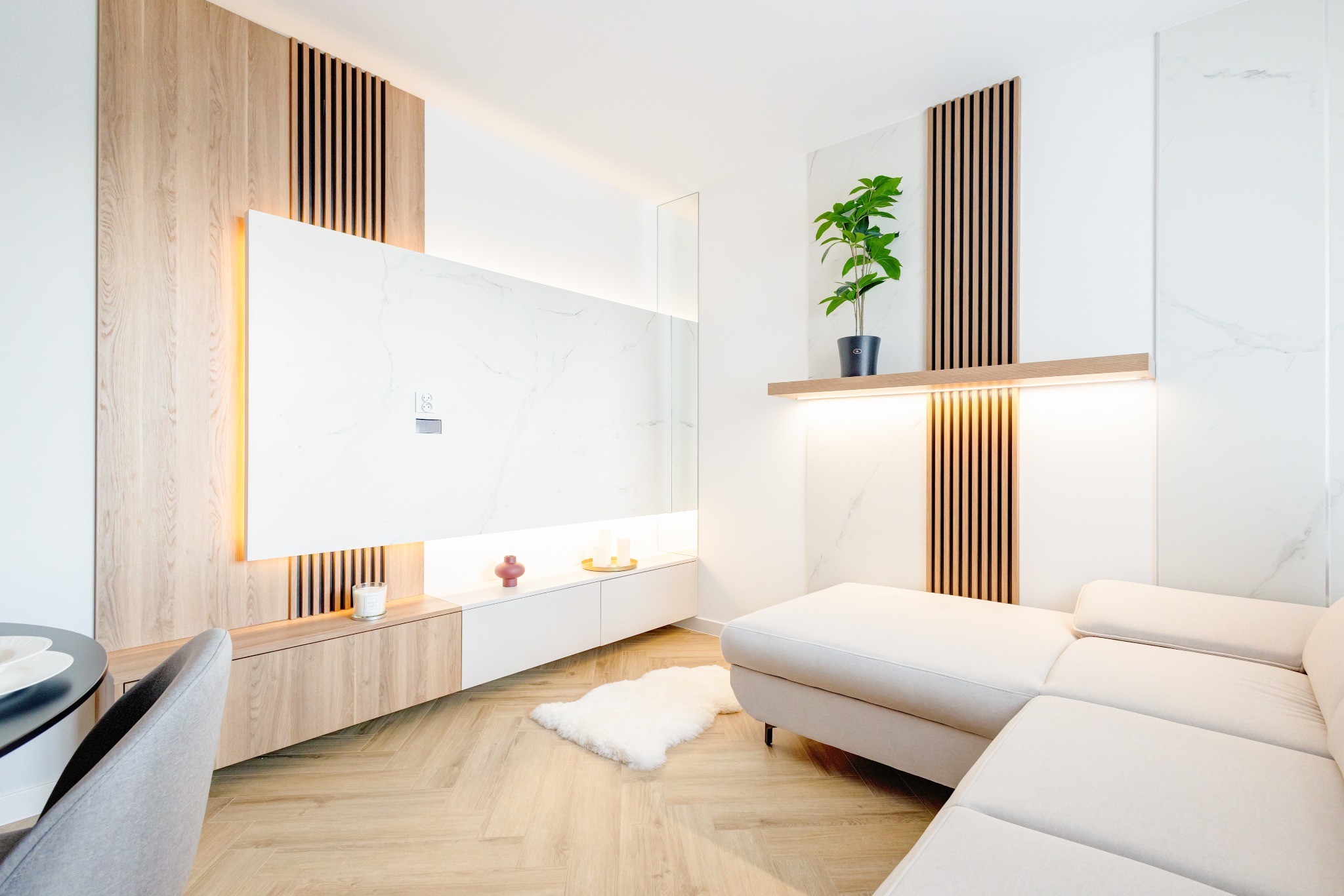 Nowoczesny salon z drewnianymi lamelami, białymi panelami imitującymi marmur i parkietem w jodełkę. Minimalistyczny design z rośliną doniczkową i subtelnym oświetleniem LED.