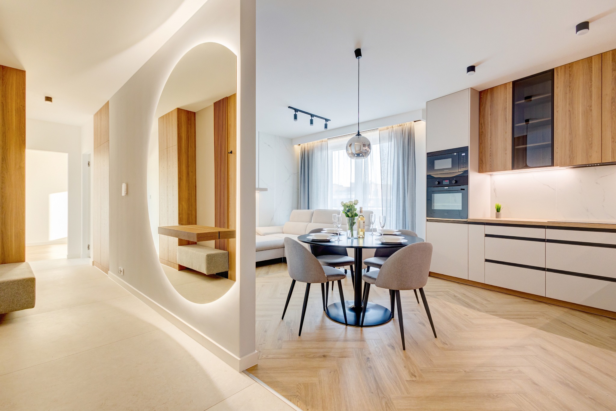 Nowoczesne wnętrze: parkiet w jodełkę, okrągłe lustro z podświetleniem LED, stół z krzesłami i zabudowa kuchenna z drewnianymi akcentami. Minimalistyczny design.