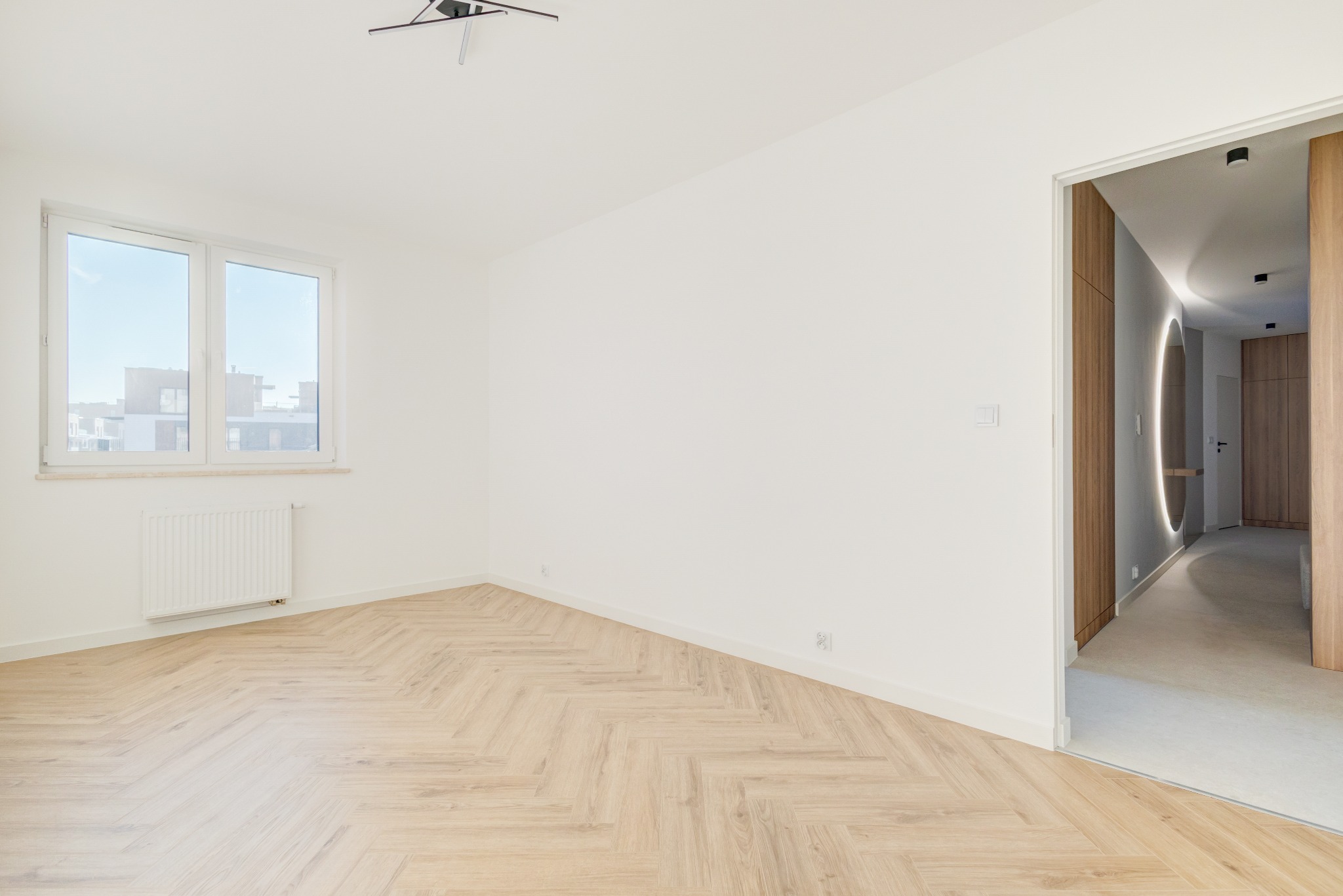 Jasne wnętrze po remoncie: parkiet w jodełkę, białe ściany, okno z widokiem na miasto i wejście do oświetlonego korytarza z drewnianymi elementami.