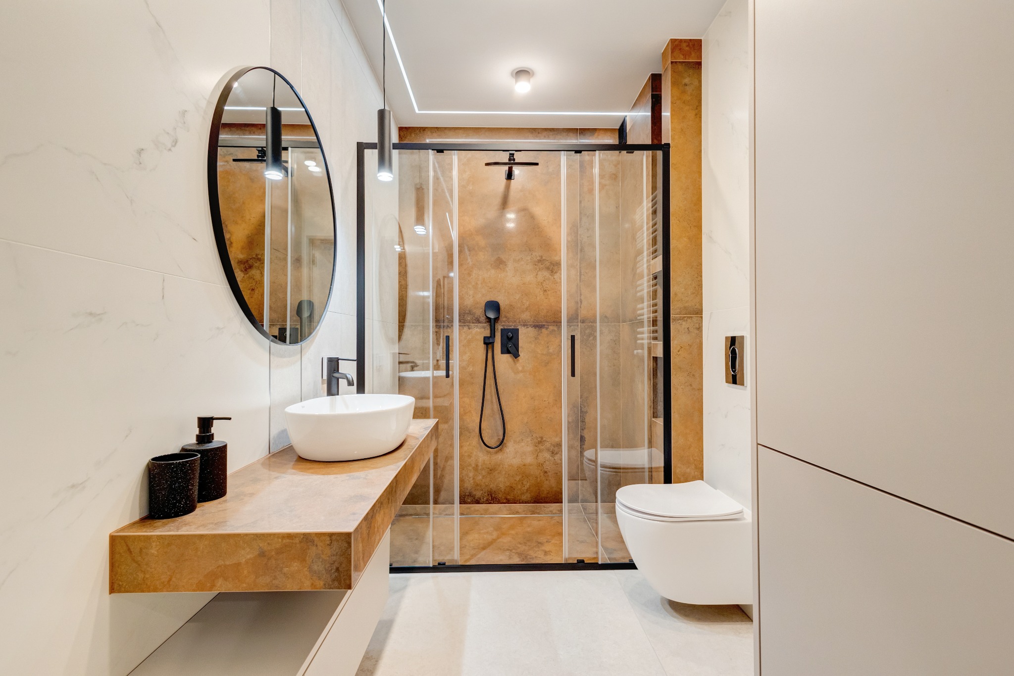 Nowoczesna łazienka z kabiną prysznicową typu walk-in, umywalką nablatową na blacie z efektem kamienia i okrągłym lustrem. Minimalistyczny design, ciepłe oświetlenie.