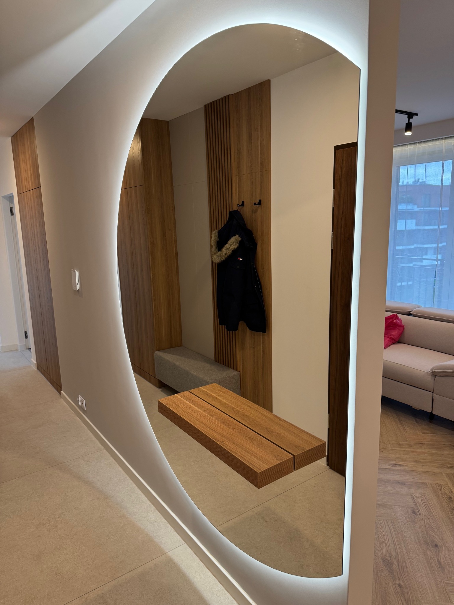 Nowoczesny przedpokój z owalnym lustrem z podświetleniem LED, drewnianą zabudową i siedziskiem. Widoczna podłoga z płytek i fragment salonu z sofą.