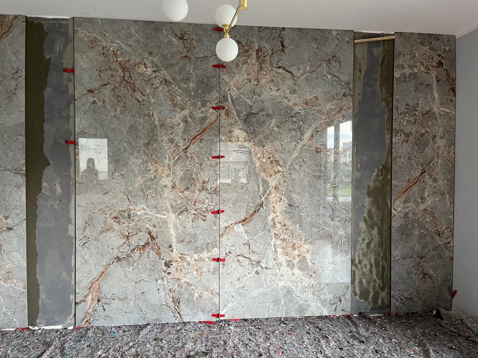 Montaż wielkoformatowych płytek imitujących marmur na ścianie, widoczne czerwone kliny poziomujące i klej. Częściowo ukończona realizacja w jasnym wnętrzu.