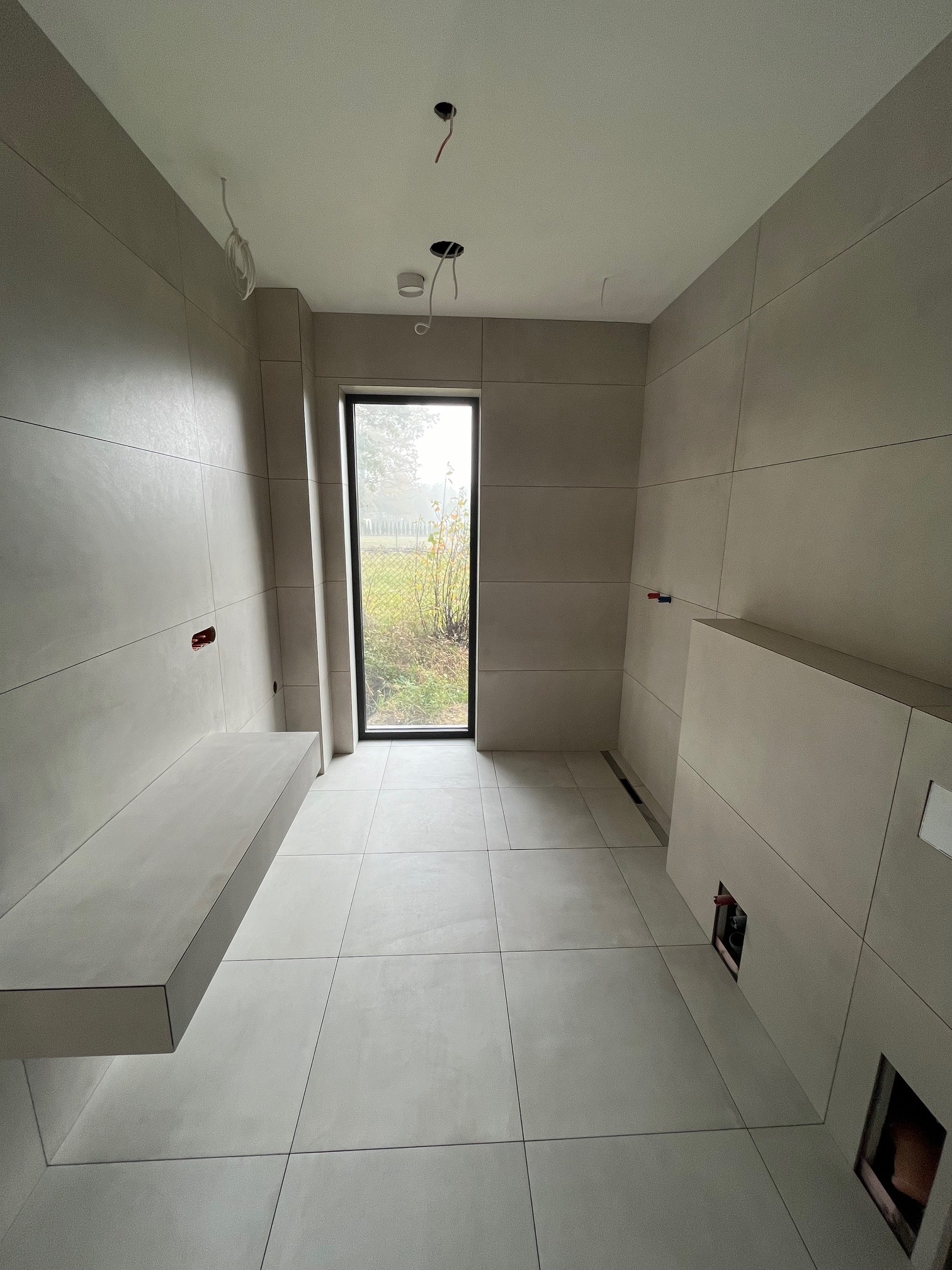 Minimalistyczna łazienka w jasnych odcieniach szarości z dużymi płytkami, oknem z widokiem na zewnątrz i elementami instalacji elektrycznej widocznymi na suficie.