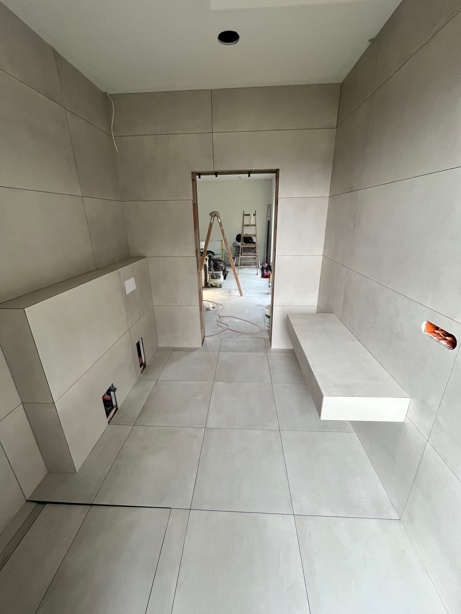 Jasna łazienka w trakcie wykańczania: płytki na ścianach i podłodze, wnęka z instalacją, półka. W tle drabina i narzędzia.