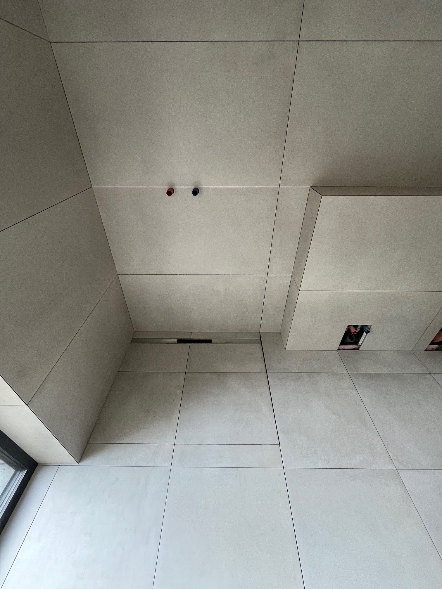 Minimalistyczne wykończenie wnętrza z szarymi, wielkoformatowymi płytami na ścianach i podłodze, z widocznymi elementami instalacji i oknem w lewym dolnym rogu.