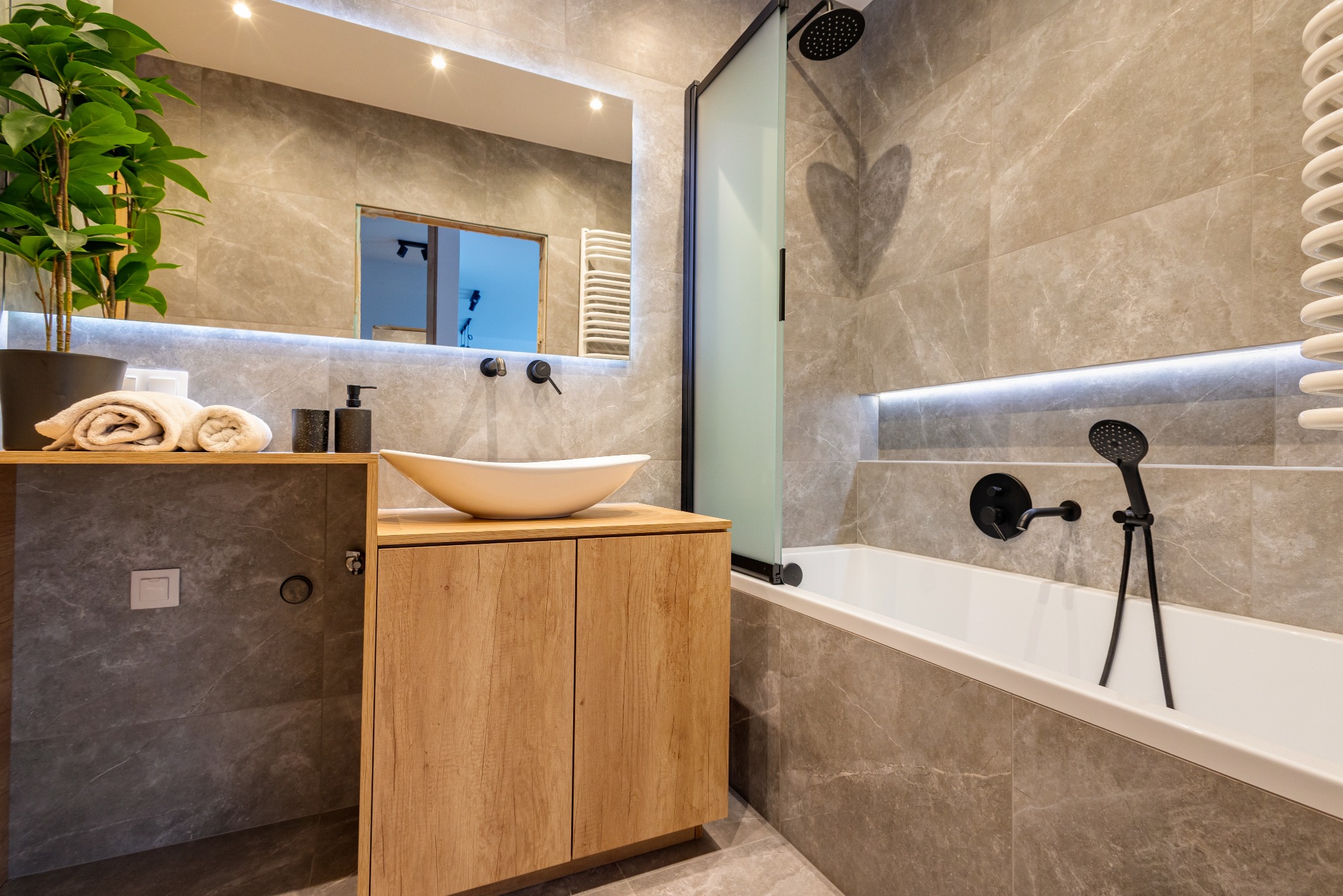 Nowoczesna łazienka z marmurowymi płytkami, drewnianą szafką pod umywalką i czarną armaturą. Oświetlenie LED podkreśla detale. Roślina w doniczce dodaje świeżości.