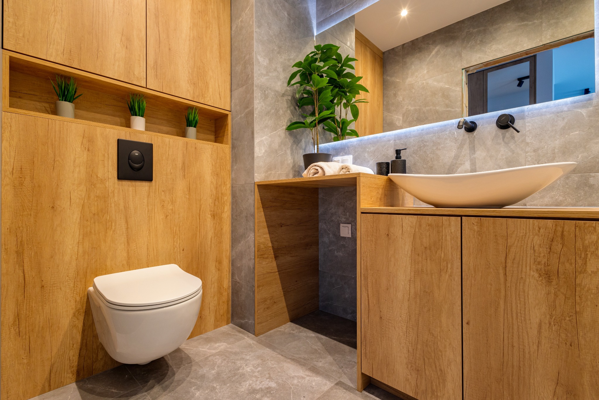 Nowoczesna łazienka z drewnianymi szafkami, szarą płytką imitującą marmur i owalną umywalką. Uwagę zwraca wisząca toaleta i roślina doniczkowa na blacie.