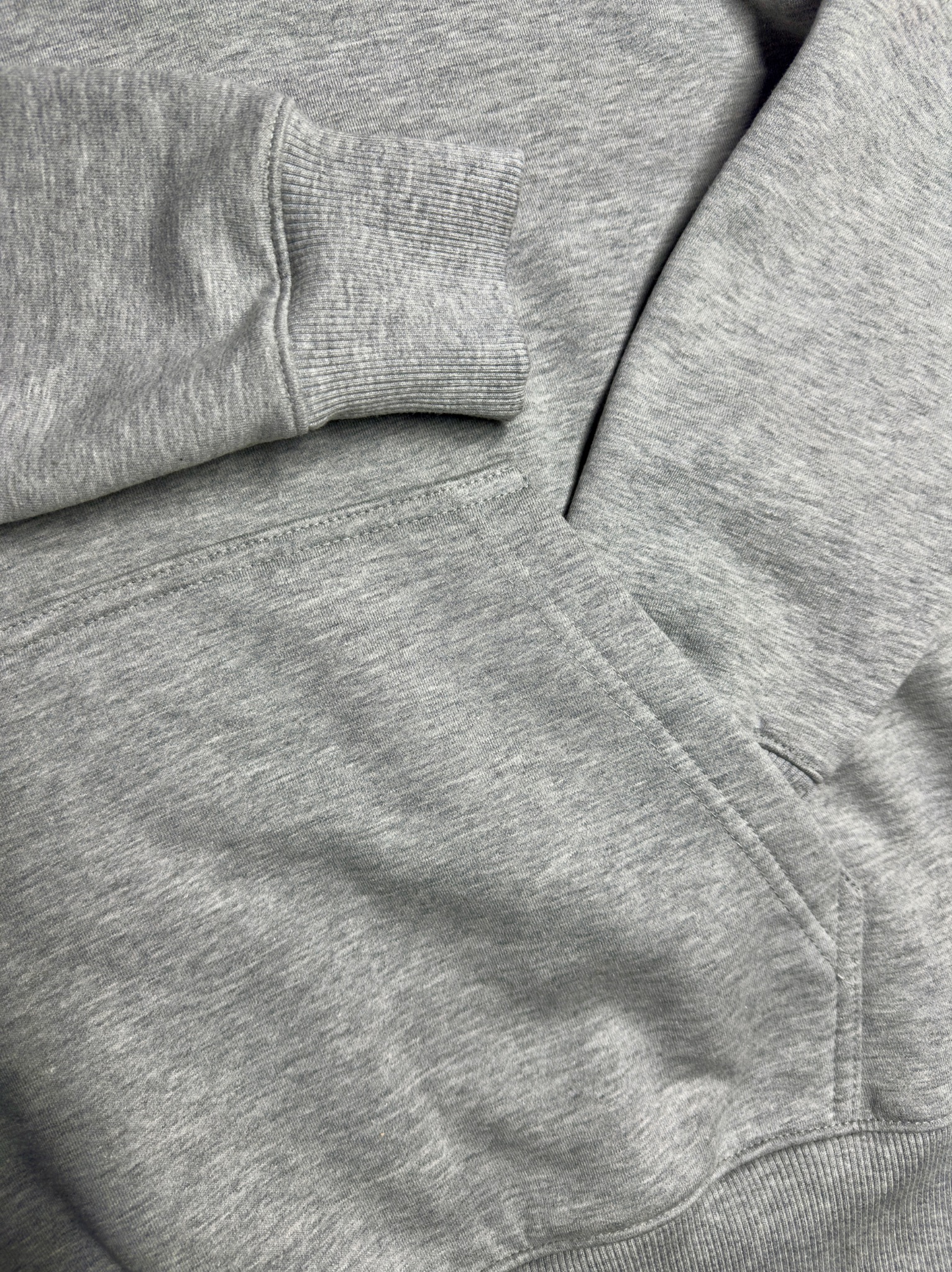 Fragment szarej bluzy dresowej z kapturem, widoczny rękaw i kieszeń, detale szycia i faktura materiału.