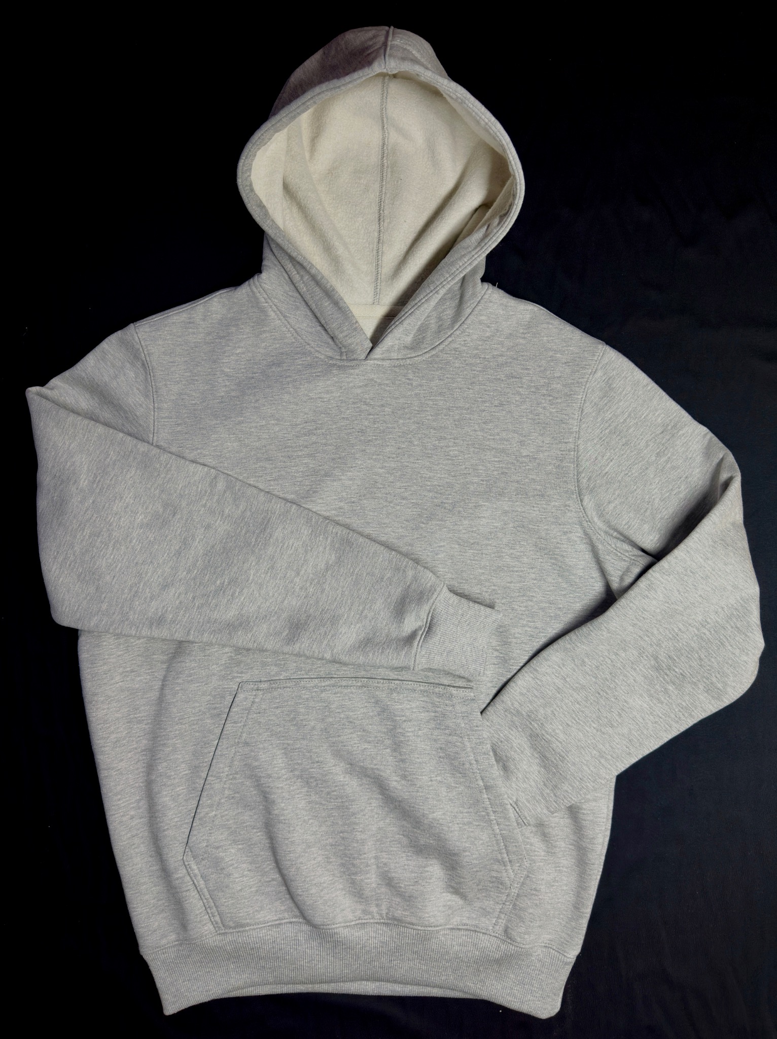 Szara bluza z kapturem typu kangurka, z jasnym wnętrzem kaptura, leżąca na czarnym tle.