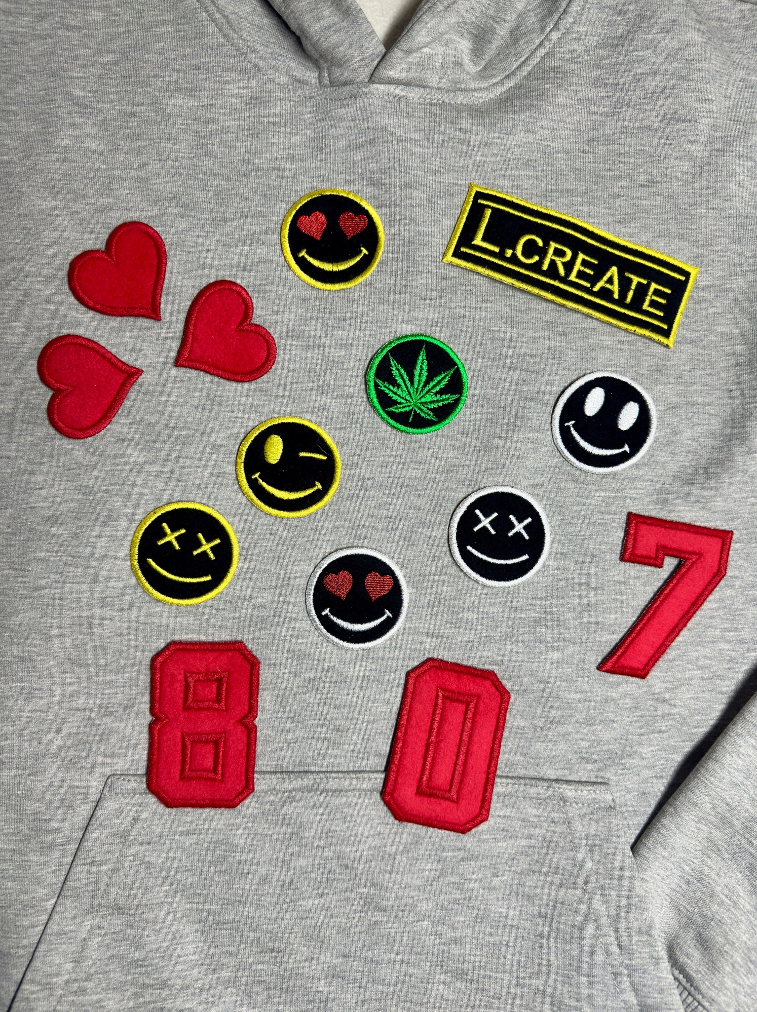 Szara bluza z kapturem ozdobiona naszywkami: emotikony, serca, liść marihuany, napis 'L.CREATE' oraz cyfry '807'.