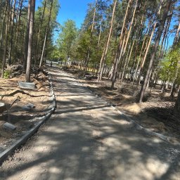 AG Ogrody I Ziemia - Budowa drogi leśnej: widok na ułożone obrzeża betonowe, przygotowany grunt, drzewa po bokach. Etap prac przed utwardzeniem nawierzchni.