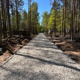 Utwardzona, żwirowa ścieżka przez las, wykończona obrzeżami. Po lewej stronie sterta ściętych pni drzew. Błękitne niebo w tle. Słoneczny dzień.