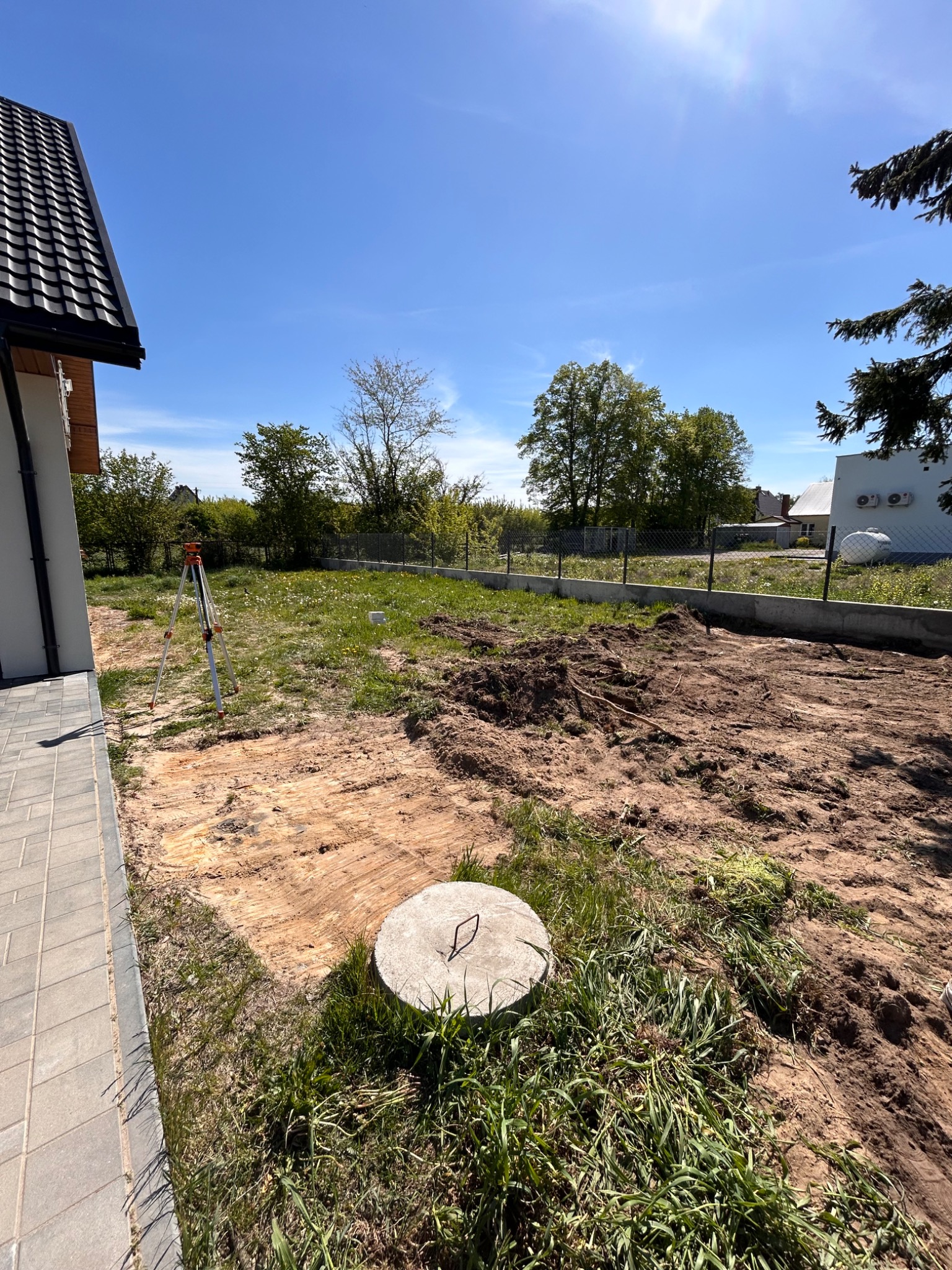 Teren po niwelacji, widoczny niwelator, częściowo usunięta darń, przygotowany pod dalsze prace ogrodowe. Betonowa studzienka w trawie.