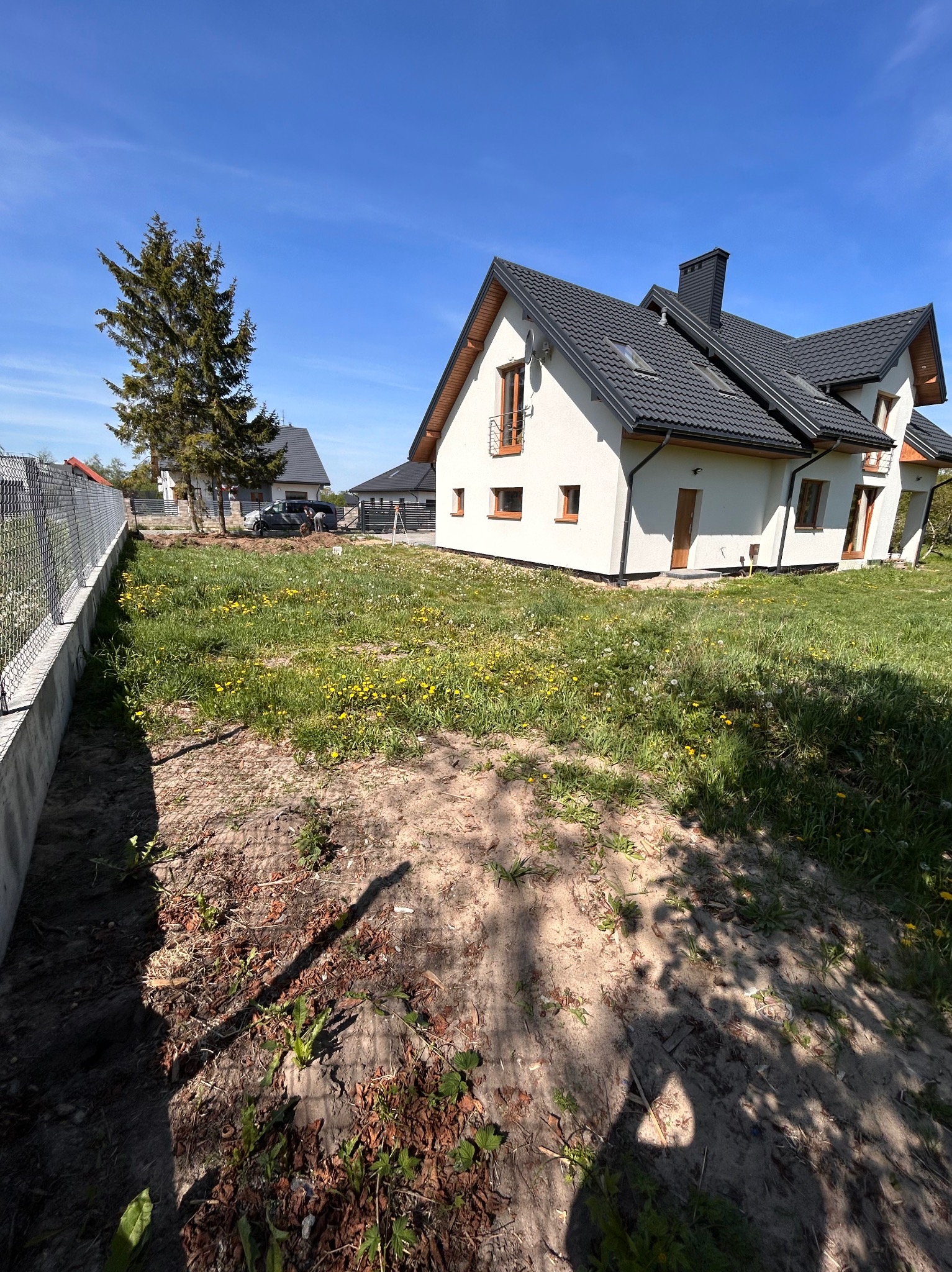 Teren przed domem z widocznym fragmentem trawnika z mleczami, obok betonowe ogrodzenie i dom z ciemnym dachem, błękitne niebo.