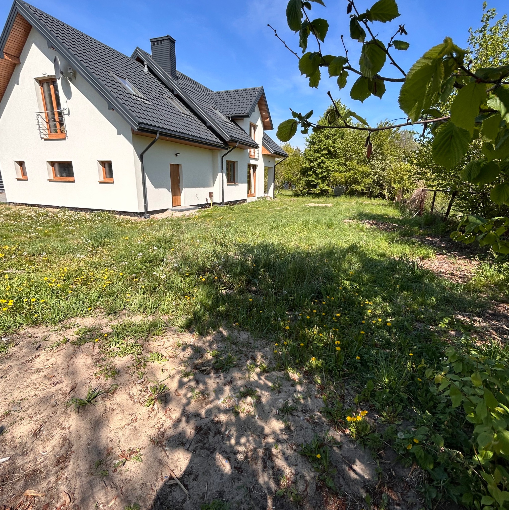 Nowoczesny dom z ciemną dachówką i jasną elewacją, otoczony trawnikiem z mleczami, widziany w słoneczny dzień z perspektywy ogrodu.